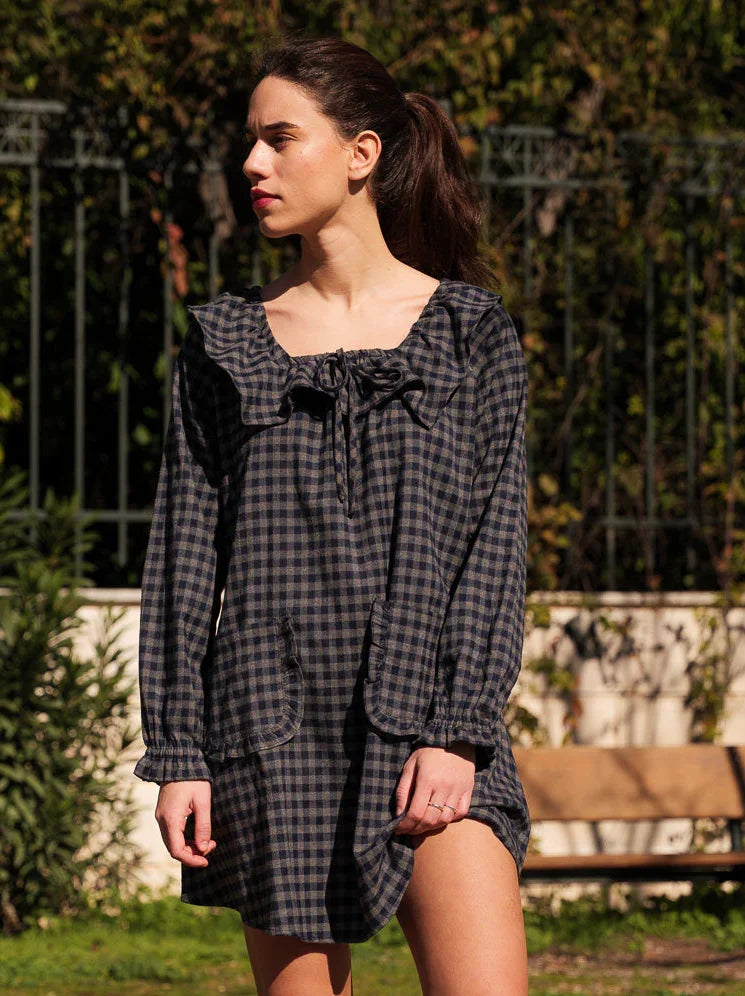 Woman in a plaid dress walking outdoors with trees and a bench in the background